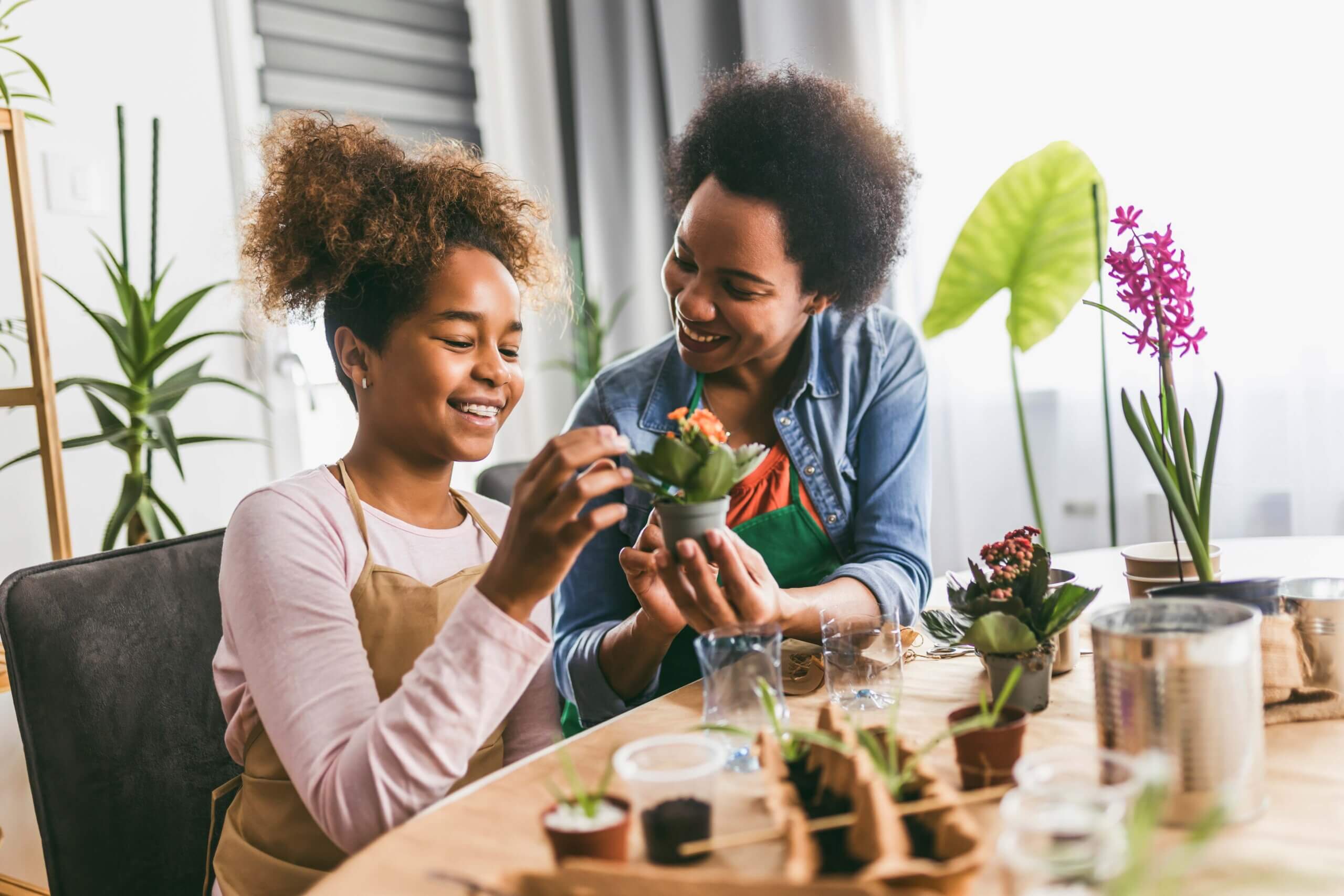 Woman with her daughter, sat at a table together as they tend to one of many mini potted plants spread out on the table's surface. The young girl is inspecting the small succulent in her hands while the mom teaches her about taking care of it. 